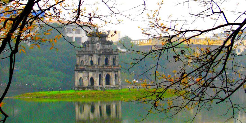 Turtle Tower on Hoàn Kiếm Lake in Hanoi, Vietnam, reflected in the water and framed by overhanging tree branches.